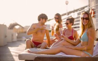 Happy friends drinking tasty beverage while sunbathing together on beach in sunny summer day